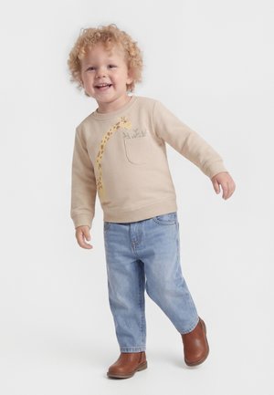 Smilende småbarn med krøllet blondt hår, iført beige sweatshirt med girafprint, lyseblå jeans og brune støvler, stående på hvid baggrund.