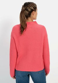 Geribbelde, oversized trui in felroze met een hoge hals. De stof heeft een zachte textuur en de achterkant heeft een iets langere zoom. Gecombineerd met blauwe jeans.