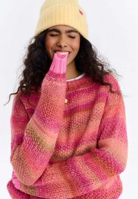 Kobieta z kręconymi włosami w żółtym kapeluszu i różowym swetrze w paski, uśmiechająca się z zamkniętymi oczami, gryząca rękaw swojego swetra.