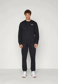 Giovane uomo con capelli ricci, che indossa una felpa nera Reebok e pantaloni, e sneakers bianche, in piedi davanti a uno sfondo chiaro e semplice.