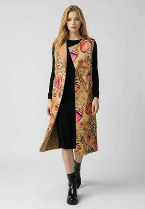 Joven mujer vestida con un vestido negro y un abrigo beige largo sin mangas con labios rosas y un patrón abstracto, combinado con botines negros.