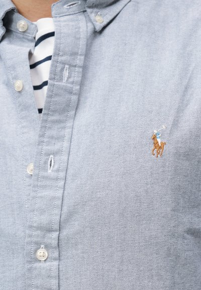 Κοντινό πλάνο ενός ανοιχτό μπλε πουκάμισου με κουμπιά, στο οποίο είναι κεντημένο το λογότυπο ενός παίκτη πόλο στο στήθος, από πάνω από ένα λευκό και σκούρο μπλε ριγέ πουκάμισο.