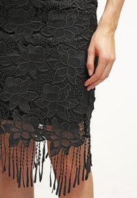 Robe noire en dentelle avec broderie florale, dotée d'un ourlet à franges. Le tissu texturé est orné de motifs floraux complexes.