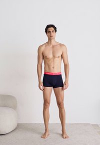 Marineblauwe boxershorts met een roze elastische tailleband waarop de merknaam staat. Het model staat blootsvoets op een neutrale achtergrond.
