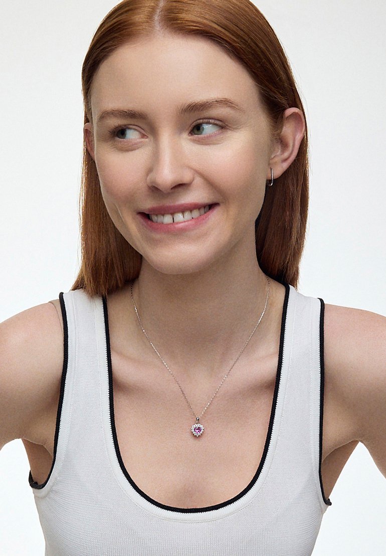 Srebrny naszyjnik w formie wisiorka z sercowym wzorem, z różowym kamieniem w centrum i białymi akcentami, noszony na jasnoszarym podkoszulku.