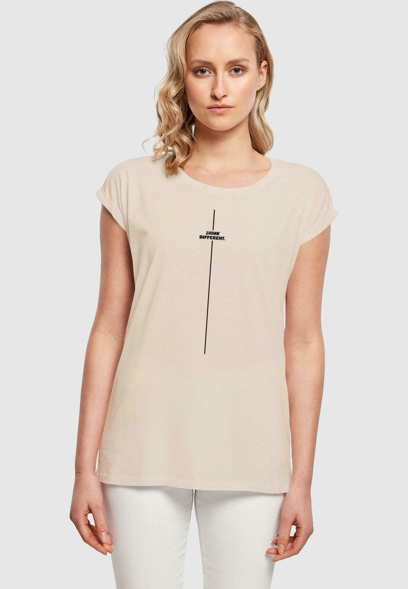Beige T-Shirt mit kurzen Ärmeln und rundem Ausschnitt, vorne bedruckt mit dem schwarzen Text "I THINK DIFFERENT.", getragen mit weißen Hosen.