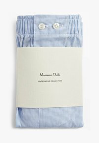 Sous-vêtements à carreaux bleus, emballés avec une étiquette de couleur crème portant le nom de la marque et le titre de la collection, maintenus par deux boutons blancs à la taille.