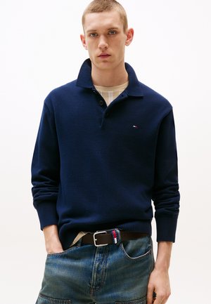 Junger Mann trägt ein marineblaues langärmeliges Poloshirt, beiges Unterhemd, braunen Gürtel mit Streifen und blaue Jeans mit der Hand in der Hosentasche.