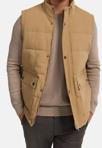Tan, mouwloze puffer vest met een hoge kraag, voorrits en twee drukknopzakken; gedragen over een lichtbruine trui.