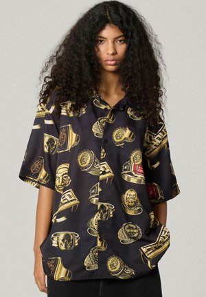 Mujer con cabello largo y rizado que lleva una camiseta negra de manga corta con diseños gráficos de anillos dorados y rojos, manos en los bolsillos, fondo neutro.