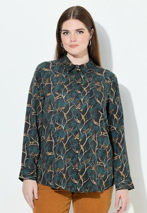 Eine Frau mit langen braunen Haaren trägt Creolen, ein dunkles Hemd mit Knöpfen, das ein Muster aus Vögeln und Bäumen zeigt, und senfgelbe Hosen. Sie steht vor einem weißen Hintergrund.