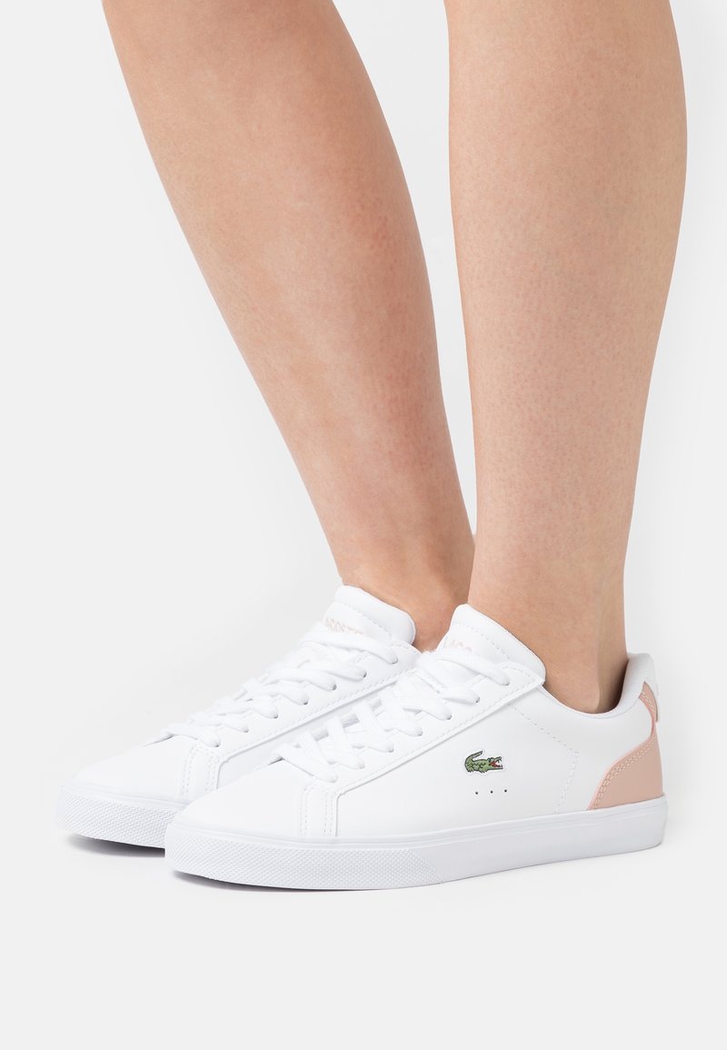 Baskets blanches à lacets en matière synthétique, avec un accent rose au talon et un logo vert. Texture lisse, bout arrondi.