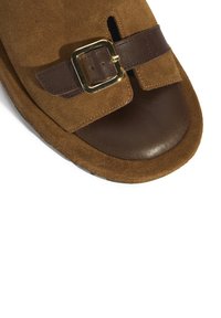 Braune Wildledersandale mit einem braunen Lederband und einer goldenen Metallschnalle sowie einem offenen Zehenbereich. Strukturierte Sohle mit abgerundeten Kanten.