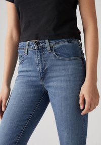 Jean skinny en denim bleu clair avec une texture lisse, poches avant et fermeture à bouton, associé à un T-shirt noir court.