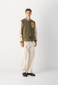 Veste bomber vert olive et beige avec des rayures jaunes et un patch "G", associée à un pantalon crème et des chaussures noires. Texture lisse, design athlétique.