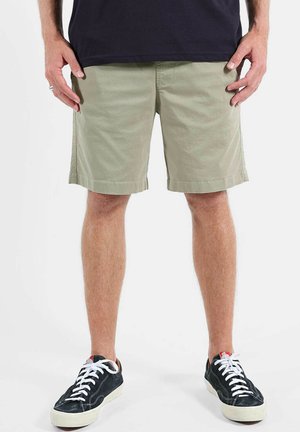 Homme portant un short kaki clair, des baskets noires avec des lacets blancs, et un t-shirt noir, debout devant un fond blanc uni.