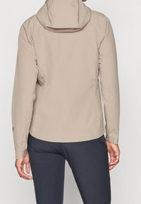 Beige huppari pitkähihaisilla hihoilla, jonka päällä on tummat housut. Kuvassa henkilö näkyy takaa vaaleaa taustaa vasten.