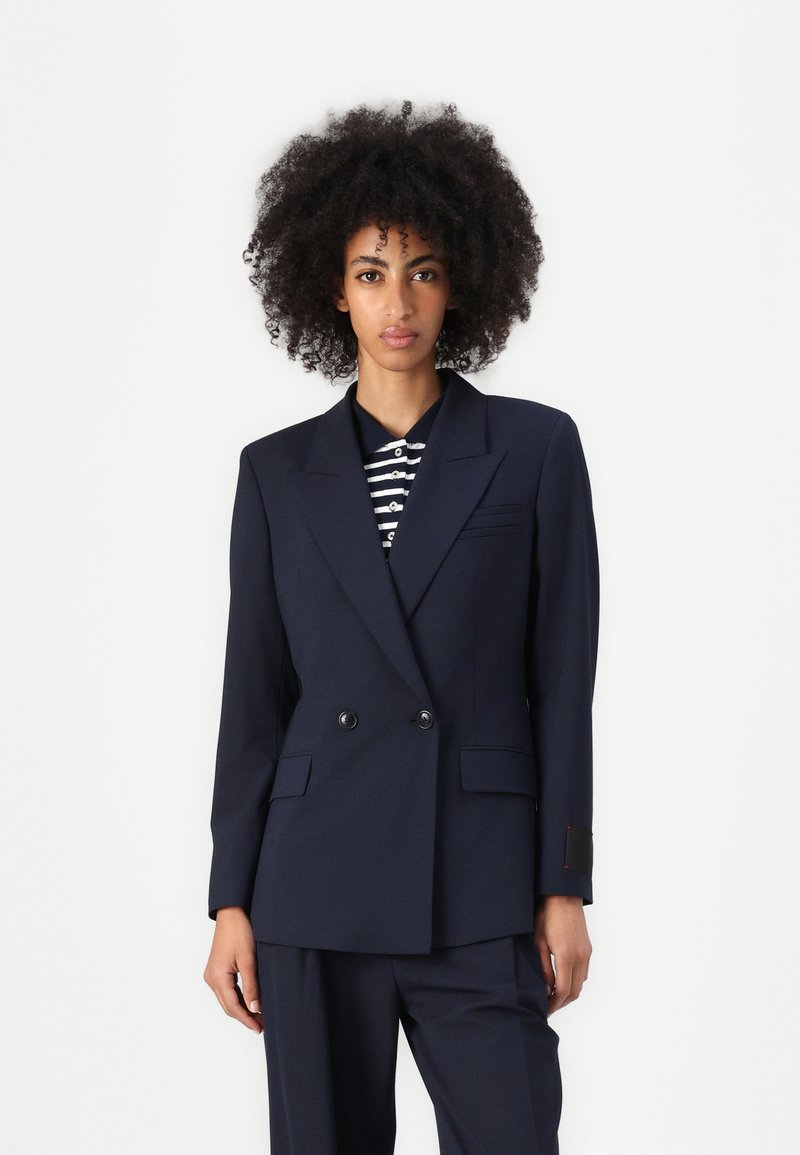 Marineblauwe dubbel-gebreide blazer met een gestructureerd ontwerp, met geknipte revers en twee voorzakken; gecombineerd met een gestreept shirt eronder.