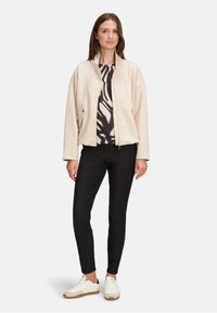 Jasno beżowa kurtka z zamkiem z przodu i bocznymi kieszeniami, noszona na czarnej i kremowej wzorzystej bluzce, połączona z czarnymi spodniami i białymi sneakersami.