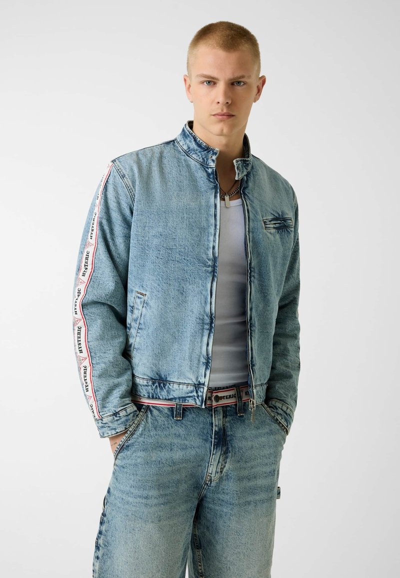 Jeune homme aux cheveux blonds courts portant une veste en jean bleu clair et un jean, un débardeur blanc et une chaîne autour du cou, les mains dans les poches.