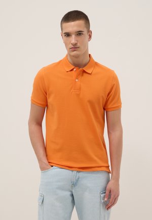 Jonge man met kort haar die een oranje polo met korte mouwen en lichtblauwe cargobroek draagt, staand tegen een effen achtergrond.