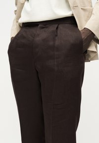 Κοντινό πλάνο προσώπου που φορά καφέ σκούρο παντελόνι με πλαϊνές τσέπες και μπεζ σακάκι, με τα χέρια μέσα στις τσέπες.