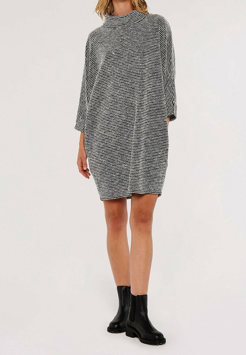 Tunique en tricot noir et blanc oversize avec col montant, manches trois-quarts et motif texturé, associée à des bottines noires à la cheville.