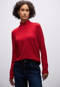 Roter Pullover mit hohem geripptem Kragen, langen Ärmeln und glatter Textur, kombiniert mit dunkelblauen Jeans, die über Fronttaschen verfügen.