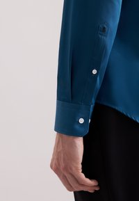 Turquoise langekoushemd met een gladde textuur. Heeft witte knopen op de manchetten en een geborduurde rozendetail op de mouw.