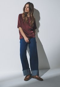 Bordeauxrotes Satintop mit kurzen Ärmeln, kombiniert mit weitem blauen Jeansstoff mit umgeschlagenen Säumen und braunen flachen Schuhen.