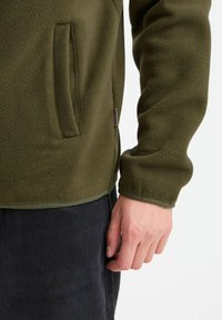 Olive green fleece jacket featuring a side pocket, ribbed cuffs, and a hem. The fabric has a soft texture and a subtle sheen.