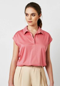 Kurzärmliges, geknöpftes Poloshirt in Korallenpink, aus glattem Stoff, kombiniert mit beigen, gefalteten Hosen.