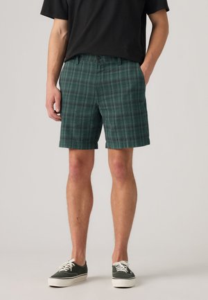 Uomo con pantaloncini verdi a quadri, maglietta nera e sneaker verde scuro con suole e lacci bianchi, in piedi su sfondo chiaro.