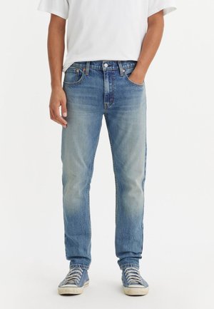 Homme portant un jean bleu clair, un t-shirt blanc et des baskets bleues, debout avec une main dans la poche devant un fond blanc uni.