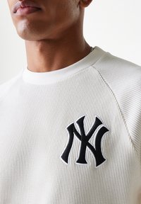 White ribbed sweatshirt featuring a black embroidered NY logo on the chest. Round neckline and textured fabric with a fitted design.