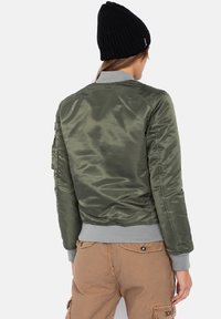 Olivgrüne Satin-Bomberjacke mit grauem Kragen und Bündchen. Sie verfügt über gerippte Säume und eine Utility-Tasche am linken Ärmel.