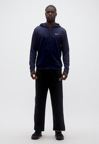 Νεαρός άντρας όρθιος, φοράει μπλε σκούρα φούτερ με φερμουάρ και λευκό λογότυπο, μαύρο άνετο παντελόνι και μαύρα αθλητικά παπούτσια σε απλό ανοιχτό φόντο.