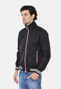 Giacca leggera nera con colletto rialzato, dotata di strisce rosse e bianche lungo la zip e sui polsini, e due tasche laterali con zip.