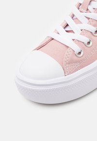 Różowy płócienny trampki z białym przodem, białymi sznurowadłami i teksturowaną białą podeszwą na jednolitym jasnym tle.