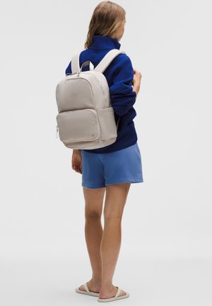 Osoba v modré bundě, modrých kraťasech a béžových sandálech, nesoucí béžový batoh, stojící zády k nám na jednobarevném pozadí.