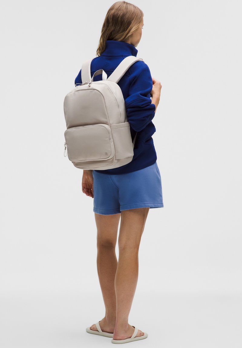 Person iført en blå jakke, blå shorts og beige sandaler, der bærer en beige rygsæk og vender væk på en ensfarvet baggrund.