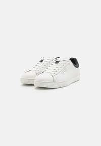 Sneaker in pelle bianca con lacci piatti, linguetta nera sul tallone, suola bianca testurizzata e branding argentato sul lato. Design classico a bassa altezza.