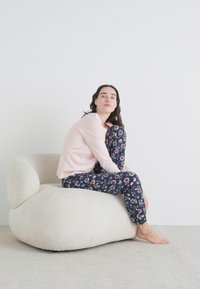 Ljusrosa långärmad topp ihop med navyblå, blommönstrade byxor. Sitter på en beige, texturerad, rund stol mot en enfärgad bakgrund.