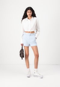 Camicia bianca corta, pantaloni corti azzurri a vita alta con cintura e sneakers bianchi. Modella che tiene una borsa scura. Tessuto liscio, stile casual.