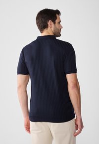 Polo cu dungi navy, cu guler, mâneci scurte, realizat dintr-un material texturat și cu un model subtil.