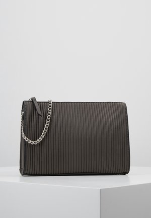 Pochette rectangulaire noire côtelée avec une chaîne argentée et un tire-zip, posée sur un cube blanc devant un fond gris clair.