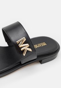 Sandale slide en cuir noir avec logo "MK" doré sur la bride et marquage "Michael Kors" doré sur la semelle, design à semelle plate.