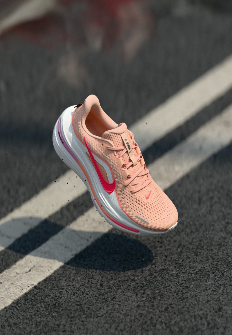 Šviesiai rožiniai Nike bėgimo batai su raudonu Swoosh logotipu ore virš asfalto ir balto kelio žymėjimo linijos.