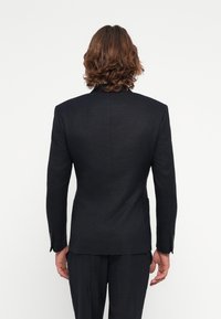 Chaqueta de traje azul marino oscuro con un tejido texturizado, abertura sencilla en la parte posterior y diseño ajustado. Presenta puños con botones y sin bolsillos visibles.