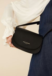 Sac bandoulière noir en cuir avec fermeture à rabat, présentant un détail de logo doré. Texture lisse, design minimaliste et bandoulière réglable.